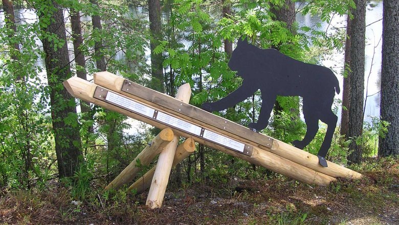 En skulptur av ett lodjur i naturlig storlek står på en piedestal av trä i skogsbrynet. I bakgrunden finns en sjö.